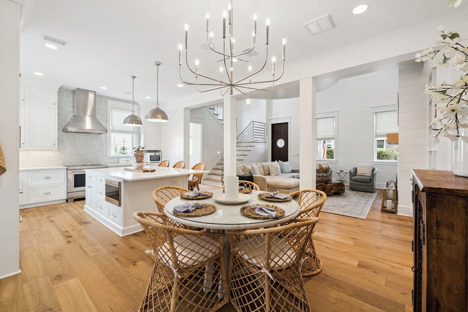 Step into your bright, open-concept sanctuary where gleaming white kitchen flows into welcoming dining space with woven chairs and modern chandelier overhead.