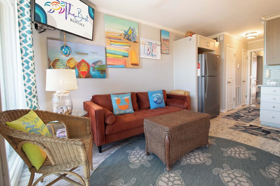 Step into your coastal retreat where wicker chairs and a cozy rust sofa invite you to unwind. Vibrant beach artwork and playful accents set a breezy, relaxed tone for your stay.