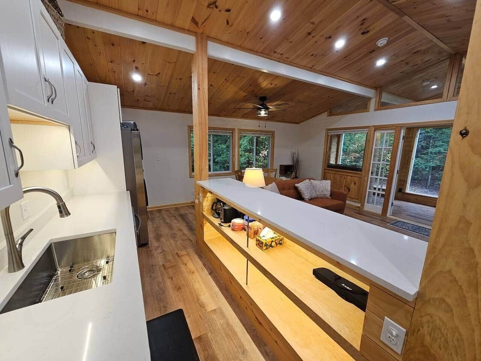 Sleek kitchen with quartz counters and white cabinetry opens to a warm living space with wood ceilings, beams, and large windows. Enjoy a modern-rustic flow perfect for cooking and gathering.
