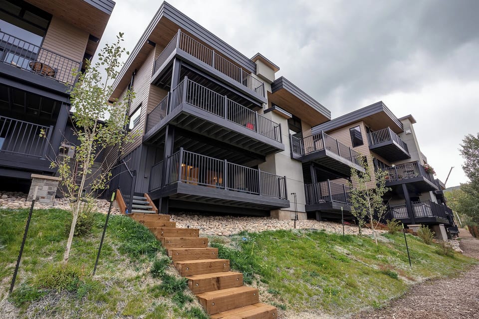 Contemporary apartment buildings with multiple balconies overlook landscaped grounds, featuring modern architecture and natural materials in a residential development.