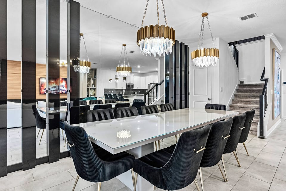 Experience our inviting dining area with a sleek table and plush black chairs, perfect for gatherings. Elegant chandeliers provide a warm glow, enhancing the modern design and showcasing the stylish kitchen.