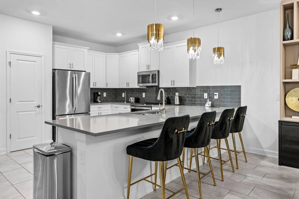 This bright, airy kitchen boasts sleek white cabinetry and a stylish gray backsplash. Gold-accented pendant lights above the island create a warm ambiance, and modern bar stools invite you to enjoy casual gatherings.