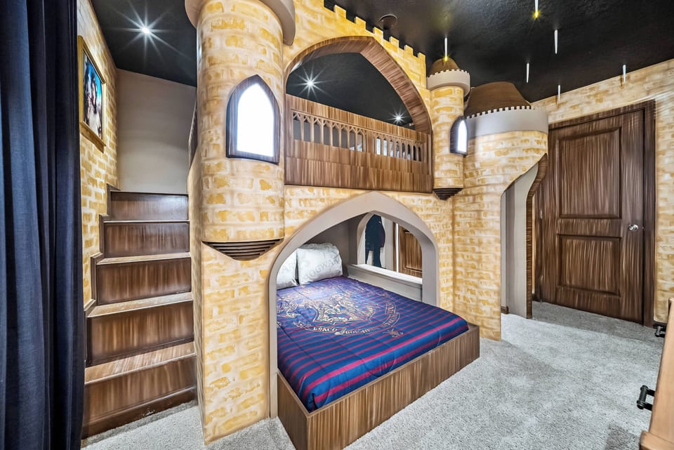 Experience a whimsical bedroom with a charming castle-inspired design, featuring a cozy bed in rich blue and red bedding. The unique arched structure and warm wooden accents create a playful, comfortable atmosphere for all ages.