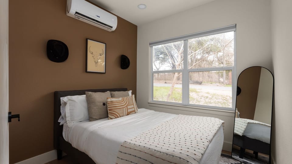 Comfortable bedroom with a full-size bed, warm accent wall, and large window for natural light. Includes a personal AC unit so you can easily control the temperature for your comfort.