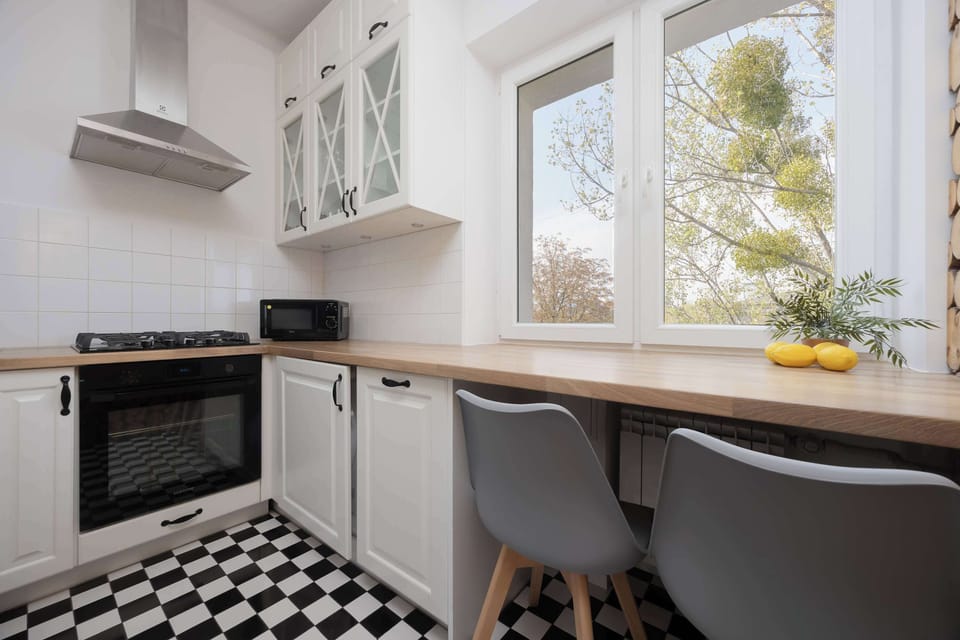 A small kitchen with white cabinets, a modern oven, and a dining area by the window. The checkered floor adds a charming retro touch.
