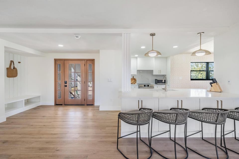 Welcoming open entryway and kitchen featuring stylish pendant lighting, rattan barstools, and warm wood tones. This bright coastal space blends modern design with laid-back charm—perfect for gathering, dining, or unwinding after a beach day.