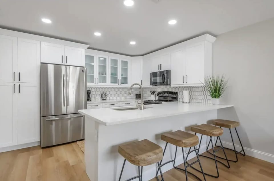 Bright and modern kitchen featuring sleek white cabinetry, stainless steel appliances, and a spacious breakfast bar with rustic stools. Perfect for cooking, dining, and entertaining in style.
