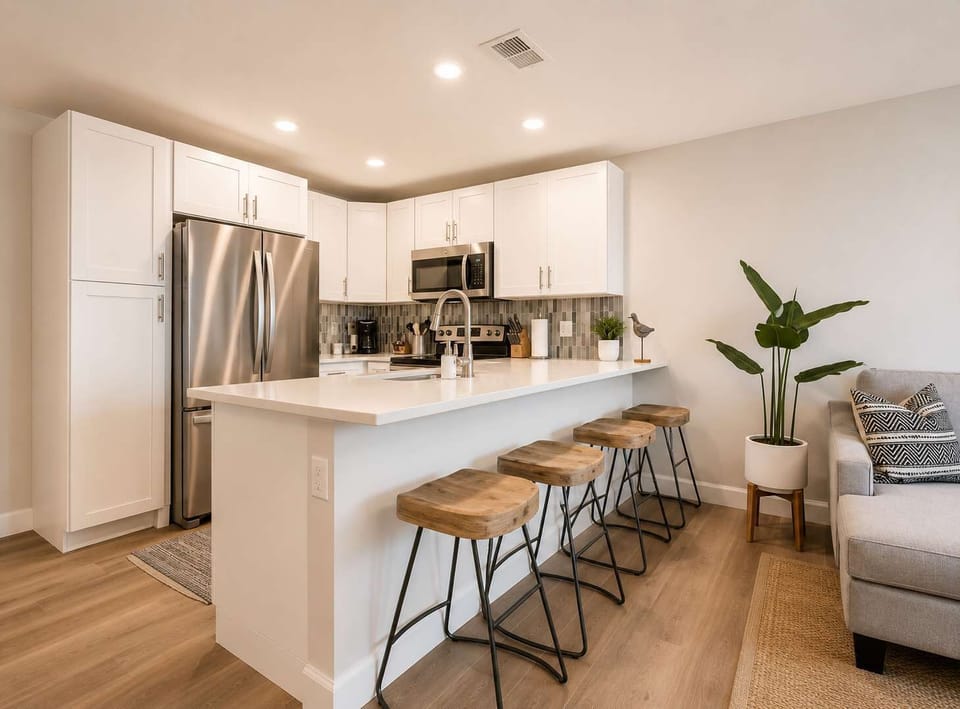 Sleek, contemporary kitchen with crisp white cabinetry, stainless steel appliances, and a spacious island with wooden bar stools. Bright lighting and modern finishes create an inviting space perfect for cooking, dining, and gathering with friends. 