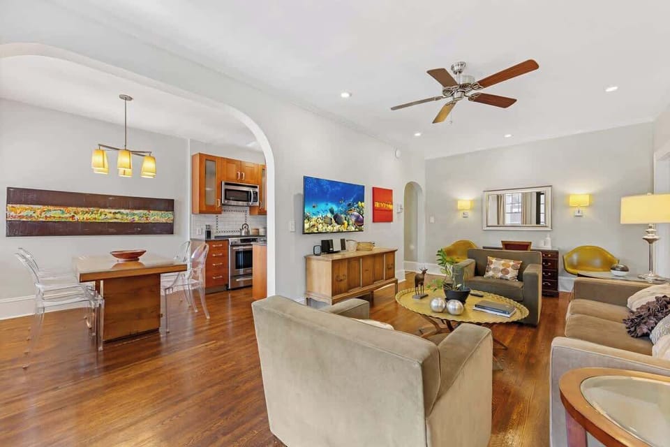 Open-concept living room in Charlotte with hardwood floors, comfortable seating, ceiling fan, and mounted smart TV. Flows seamlessly into the dining area and kitchen, creating a spacious, inviting layout for relaxing or entertaining.