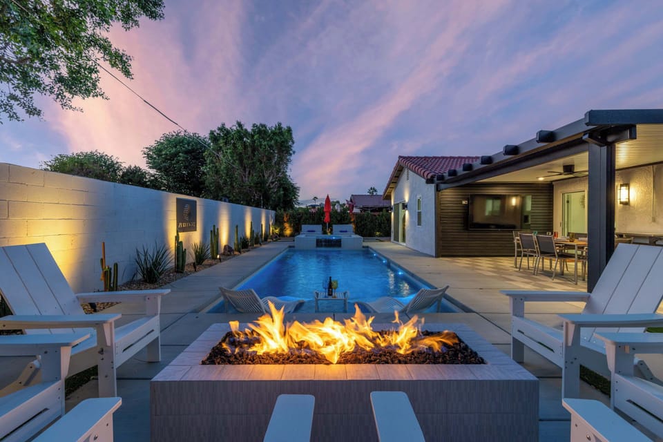 Twilight backyard — roaring gas fire pit flanked by Adirondack chairs, covered outdoor TV lounge & desert-garden accent wall