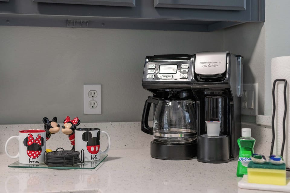 A coffee station with a dual coffee maker and themed mugs on a clean countertop.