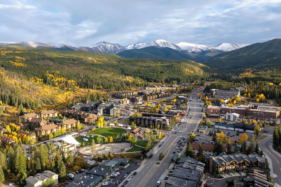 Aerial view of mountain town nestled in valley with snow-capped peaks and golden autumn foliage surrounding residential and commercial areas.