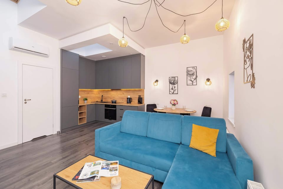 A living room area showing a blue sofa, coffee table, and kitchenette in the background. The layout emphasizes functionality and comfort.