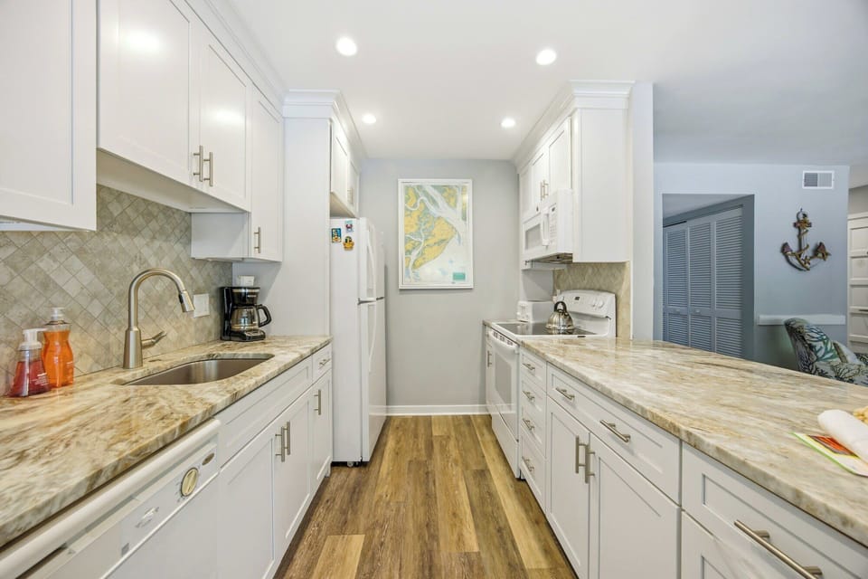 Creamy cabinetry and swirling stone counters create a crisp, airy kitchen built for slow mornings. Soft recessed lighting shines over warm wood floors, while nautical art adds a touch of coastal charm to this bright, efficient culinary space.