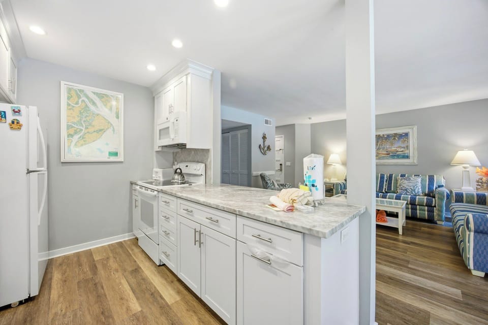 White cabinetry and stone surfaces gleam under recessed lights, creating a bright hub for vacation meals. The open layout flows seamlessly from the prep space into a lounge filled with coastal stripes and warm evening glow.