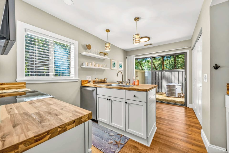 Honey-toned butcher block counters and warm wood flooring create a grounded, organic feel in this sun-drenched kitchen. Modern gold pendants glow above the island, leading your eye toward the lush greenery visible through the sliding glass doors.