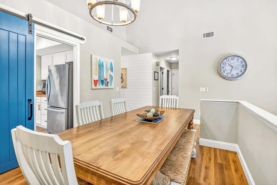 Oak grain and white slatted chairs create a coastal dining nook, anchored by a bold blue barn door and a nautical chandelier. Seagrass seating and surfboard art bring a beachy soul to this airy, open gathering place.
