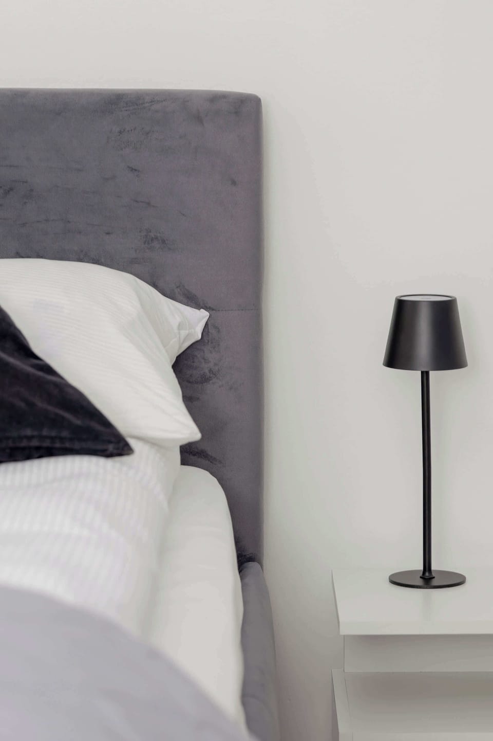 A close-up of a bedside lamp and part of a grey upholstered headboard. The setup is simple and minimalistic, emphasizing functionality.