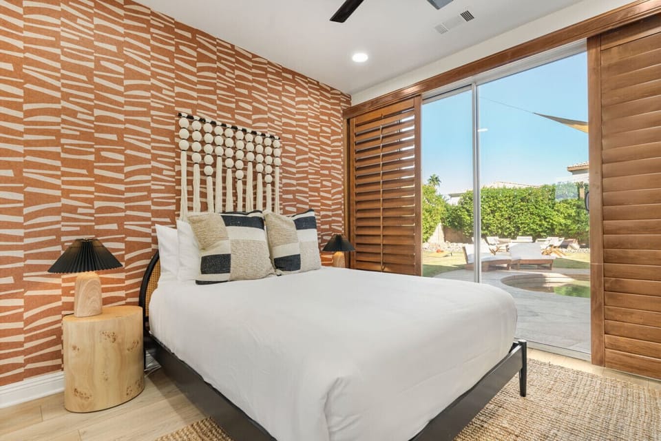 Stylish bedroom w/ textured accent wall, cozy bed, & large sliding doors opening to a sunlit outdoor lounge area.