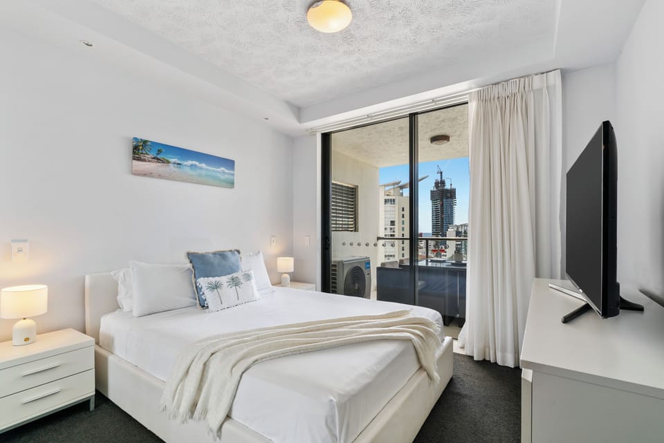 The primary bedroom is a comfortable retreat with a queen bed dressed in crisp linen, balcony access for fresh ocean air, a mirrored wardrobe, and a Smart TV for private entertainment.