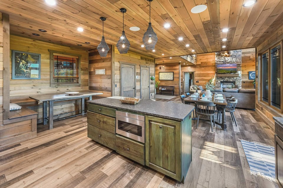 Rustic kitchen island with modern lighting opens to a cozy dining and living area with a stone fireplace.