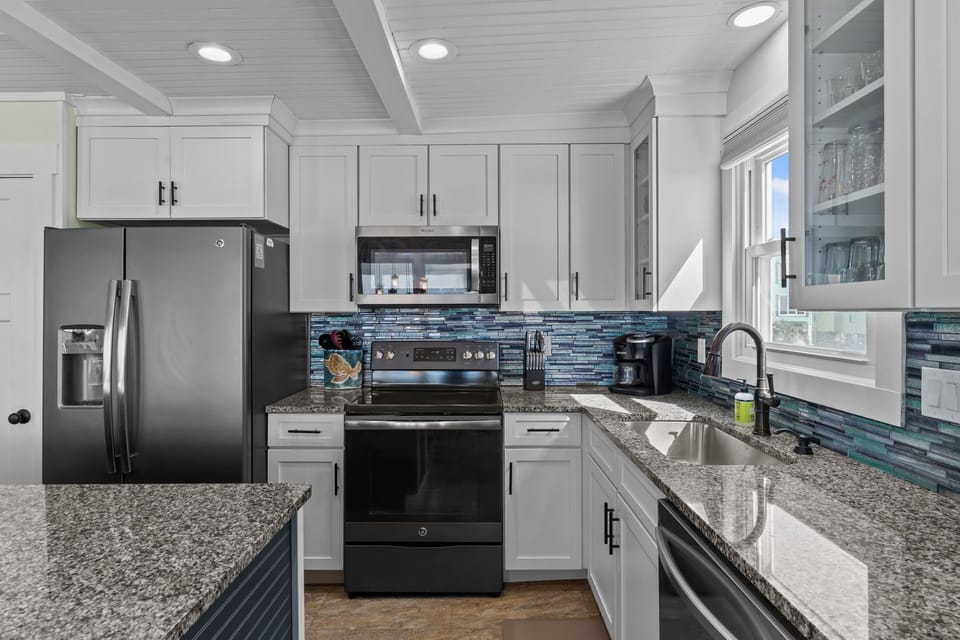hite kitchen with granite island, woven stools, stainless fridge, oven, microwave, dishwasher, tile backsplash, glass cabinets, sink window, coffee maker, knife block, shelves, and decor.