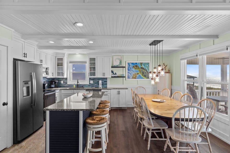 Open kitchen with island seating, granite counters, stainless appliances, tile backsplash, white cabinets, pendant lights, long dining table, chairs, wall art, and windows showing ocean and deck.