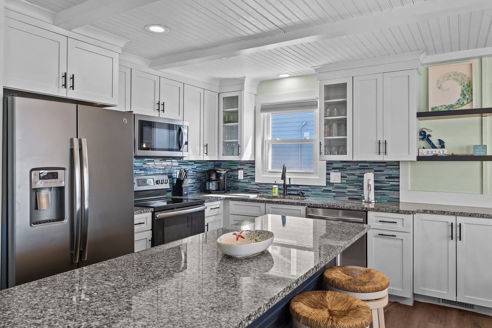 hite kitchen with granite island, woven stools, stainless fridge, oven, microwave, dishwasher, tile backsplash, glass cabinets, sink window, coffee maker, knife block, shelves, and decor.