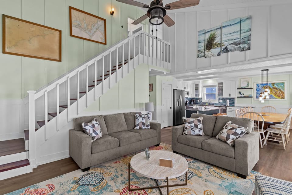 Bright living room with vaulted ceiling, fan, sofa seating, TV console, coastal art, rug, lamp, and glass door to deck; windows reveal ocean views, chairs, and “Relax Beach” sign.