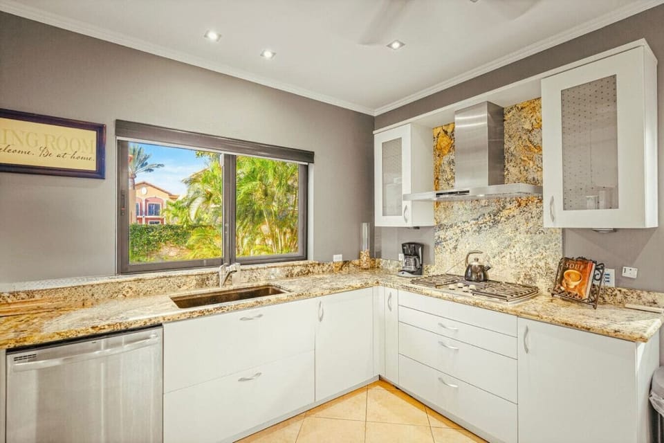 Sleek white cabinetry and golden granite glow under soft light, framed by lush garden views through wide windows.