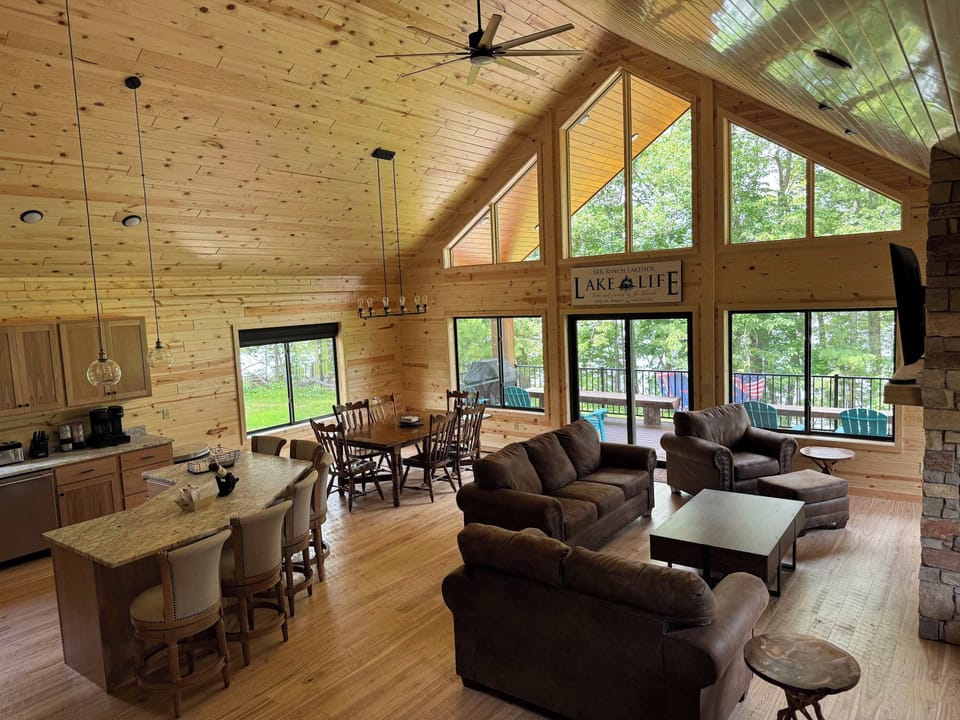 Main level entry view across Lakeside #1 great room, 6-person dining kitchen island, 6-person dining table, family room around gas fireplace, 65" smart TV, access to lake side wrap around deck with BBQ