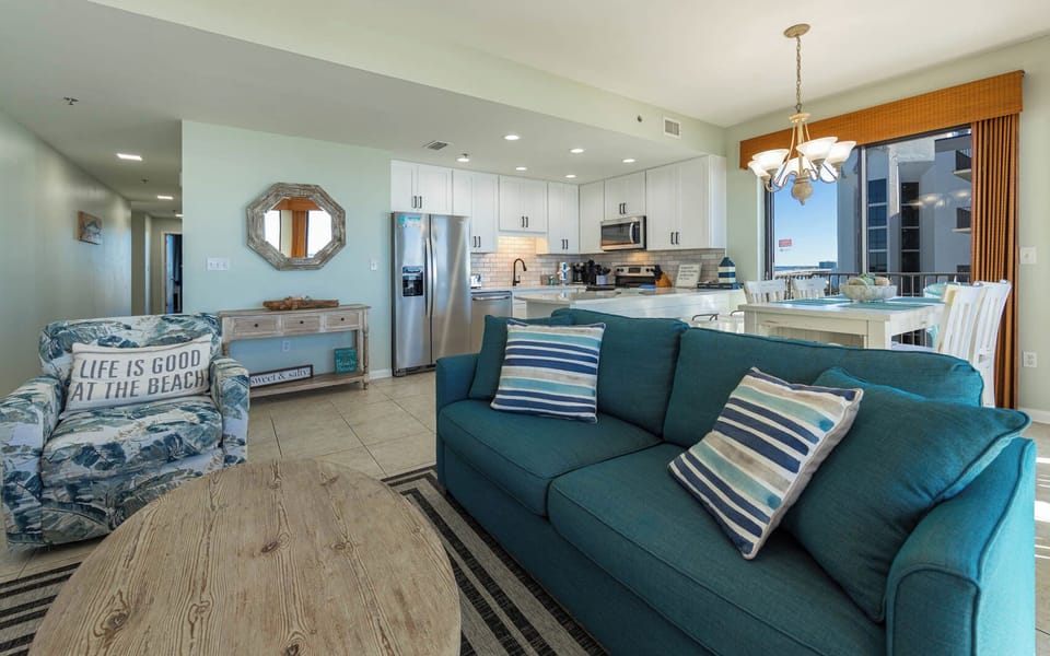 Living area features a teal sofa with striped pillows, a rustic coffee table, and a dining space with a view of the balcony