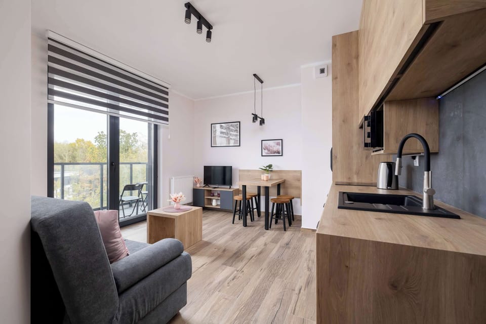 A modern living room includes a grey sofa, a coffee table, and large windows that open onto an outdoor scene.