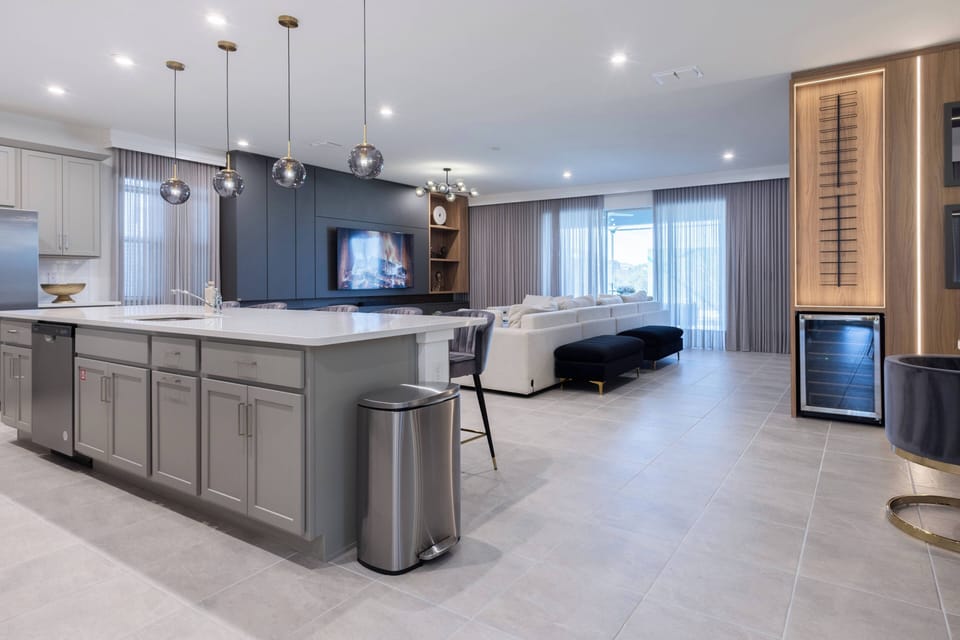 This stylish kitchen opens to the living area, featuring sleek gray cabinetry and a spacious white island perfect for casual dining. Large windows flood the space with natural light, enhancing the modern décor and warm atmosphere.