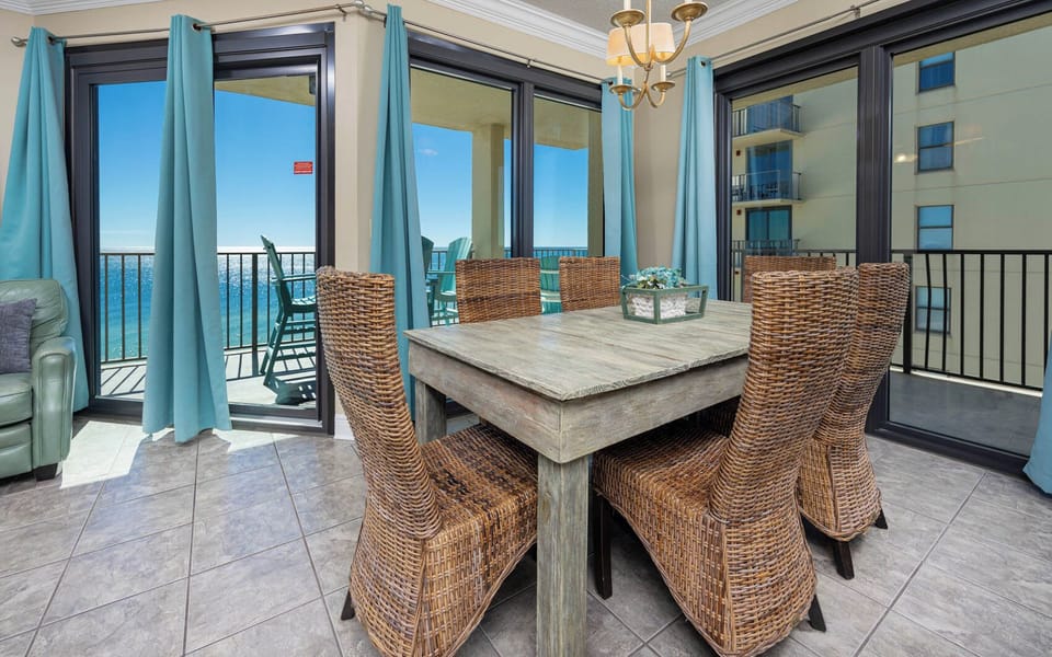 A dining area features a rustic wooden table surrounded by woven chairs, with large windows offering a beach front view