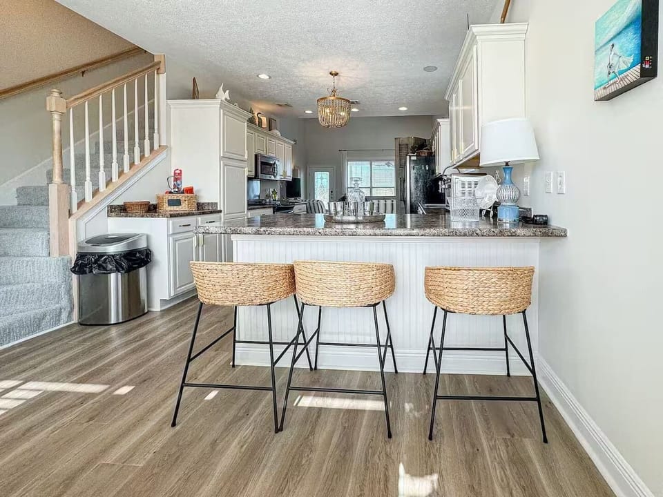 Bright, open-concept kitchen with a large island, granite countertops, and three woven bar stools. Features modern white cabinetry, wood-style flooring, and elegant lighting—perfect for cooking and gathering.