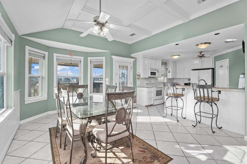 Open dining and kitchen area with glass dining table, bar seating, white cabinetry, tile floors, and bright windows.