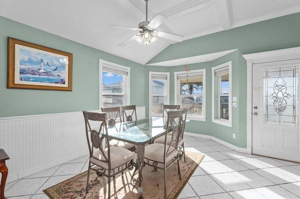 Sunny dining nook with glass table, bay windows, ceiling fan, and bright coastal decor near the home entry.