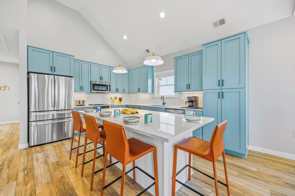 A bright, coastal-inspired kitchen invites you to gather around the spacious island, where cheerful blue cabinetry, sleek stainless steel appliances, and warm wood floors create a welcoming space for your culinary adventures.