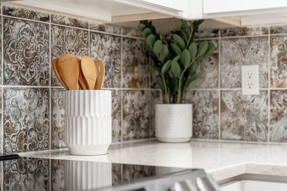 Charming kitchen details with patterned backsplash, greenery, and natural wood utensils in a modern white setting. Thoughtful touches that make this space both stylish and welcoming for every guest chef.