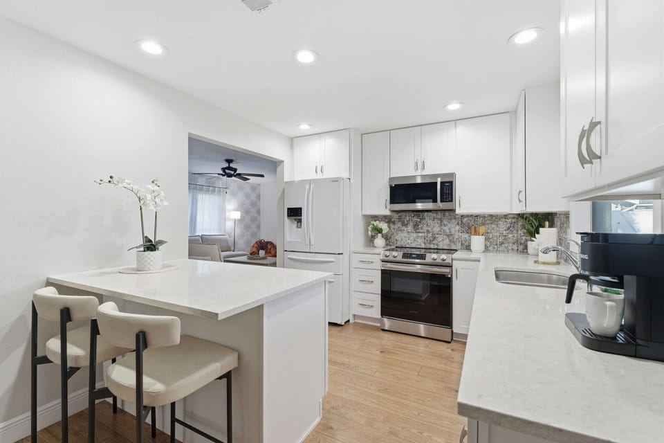 Bright and modern kitchen featuring sleek white cabinetry, stainless steel appliances, and a cozy breakfast bar. Perfectly equipped for home-cooked meals or morning coffee, blending style and functionality in every detail.