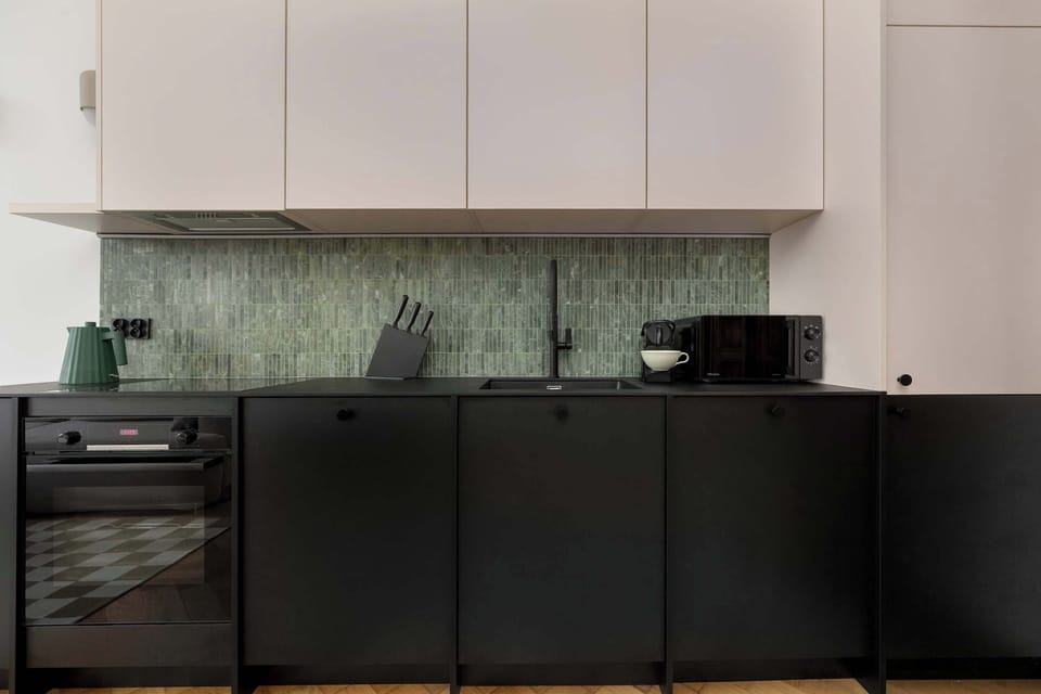 A modern kitchen with black lower cabinets and a green tile backsplash. The sleek design contrasts beautifully with the warm wooden floor.
