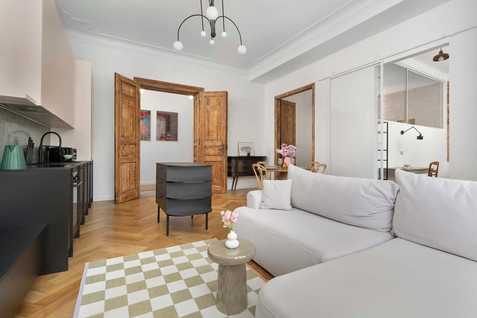 A bright living room with large windows, white walls, and a gray sofa. A patterned rug and wooden floors add warmth to the modern space.