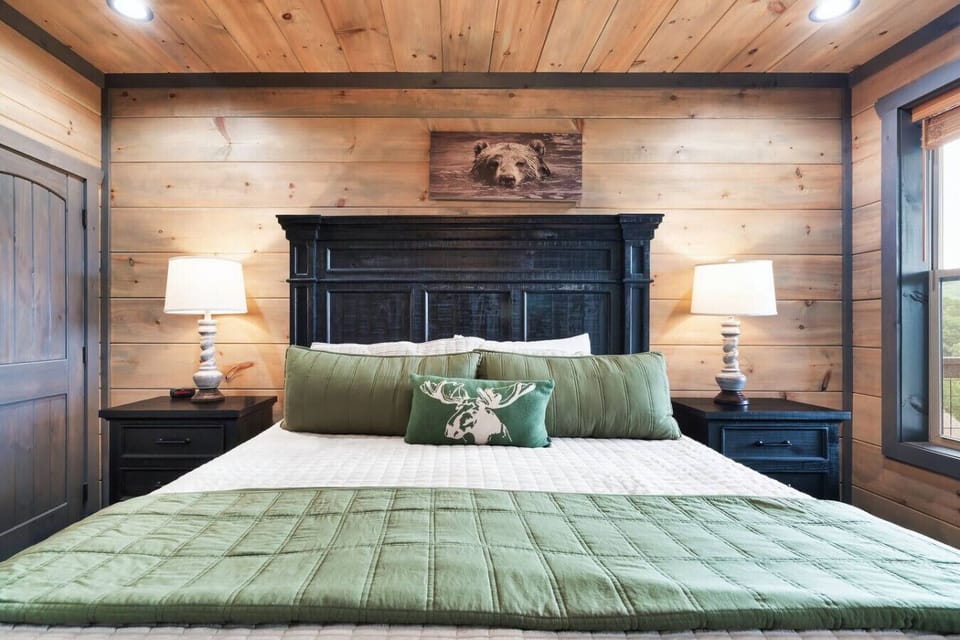 Framed by rich wood paneling and natural textures, this king suite offers a calming retreat with its deep green tones, antler pillow, and that adorable bear peeking down from above.