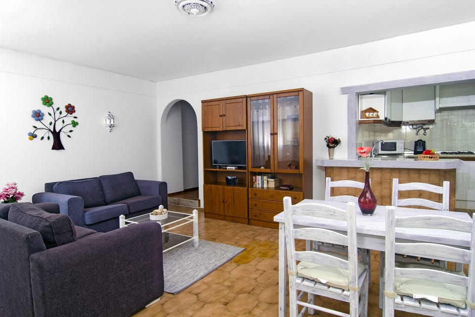 The living room is furnished with blue sofas in front of the TV and a white dining table for four people. Behind it is the passageway with counter to the open kitchen.