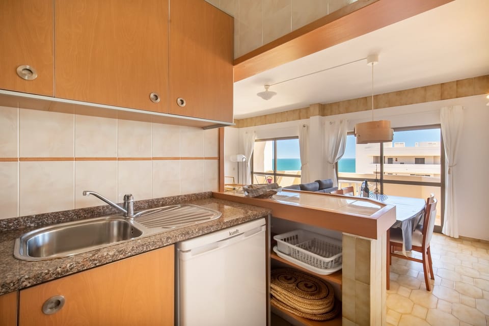 A bright, open-plan kitchenette that flows into the dining and living area, offering a superb sea view. The kitchen features light wood-toned cabinets, a dark granite-look countertop, and beige wall tiles. A small counter visually separates the space. The dining area is flooded with light from large windows overlooking the beach and the ocean.