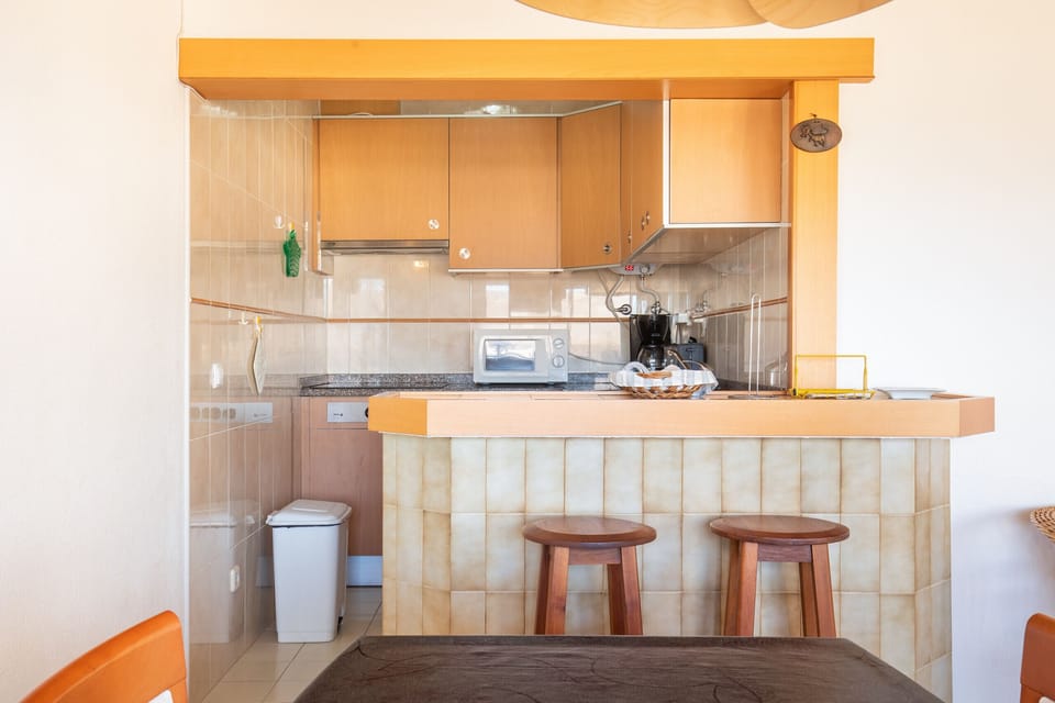A compact, bright kitchenette with a Mediterranean feel, separated by a bar with a wooden counter and two stools. The kitchen features beige wall tiles, light wood cabinets, and a bar front with larger, curved beige tiles. A microwave, coffee maker, and trash bin are visible.