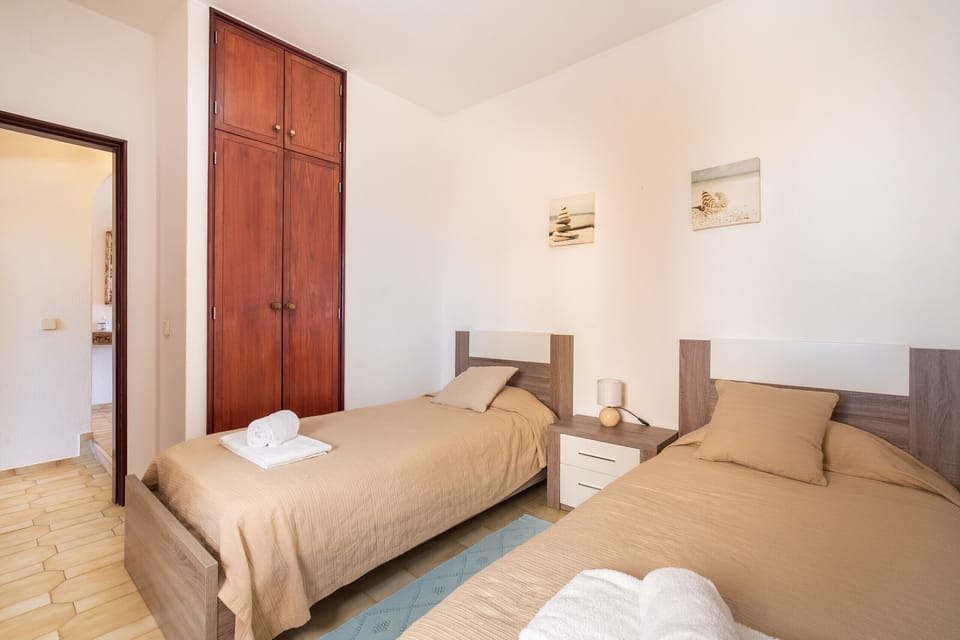 A welcoming and bright bedroom featuring two twin beds dressed in soft beige bedding. A modern nightstand with a small lamp sits between the beds. The room features a large chestnut wood wardrobe and is decorated with two framed beach-themed pictures, conveying a relaxed atmosphere.