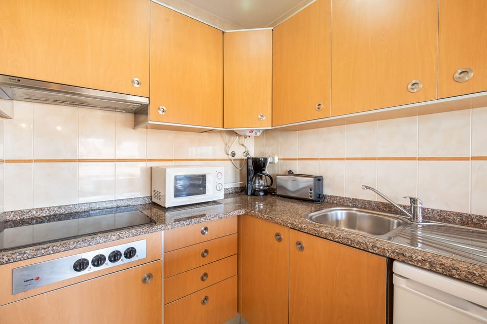 A functional, well-equipped U-shaped kitchen featuring light wood cabinetry and a dark speckled granite countertop. The walls are lined with beige tiles accented with decorative borders. The kitchen includes a ceramic hob, extractor hood, microwave, toaster, and a stainless steel sink.