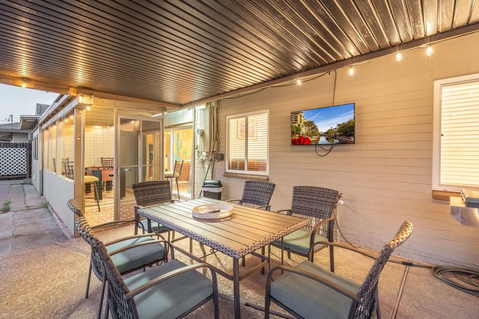 Covered patio dining area with a long table, cushioned chairs, and warm string lighting overhead—an easy spot to share meals outdoors while a wall-mounted TV adds a casual backdrop for evening hangouts.